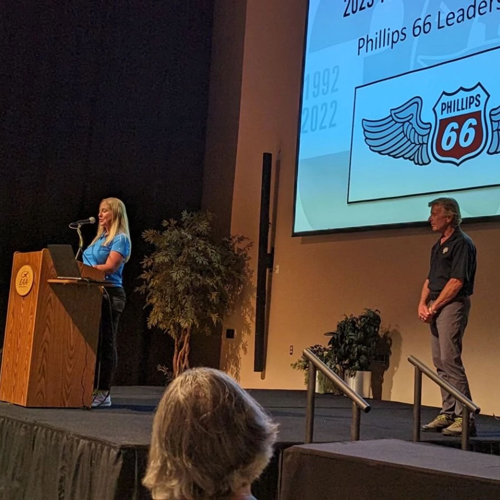 A woman speaking at a podium with a Phillips 66 Aviation logo on a screen behind her, and a man standing nearby, representing Phillips 66's community involvement and philanthropic initiatives in aviation.