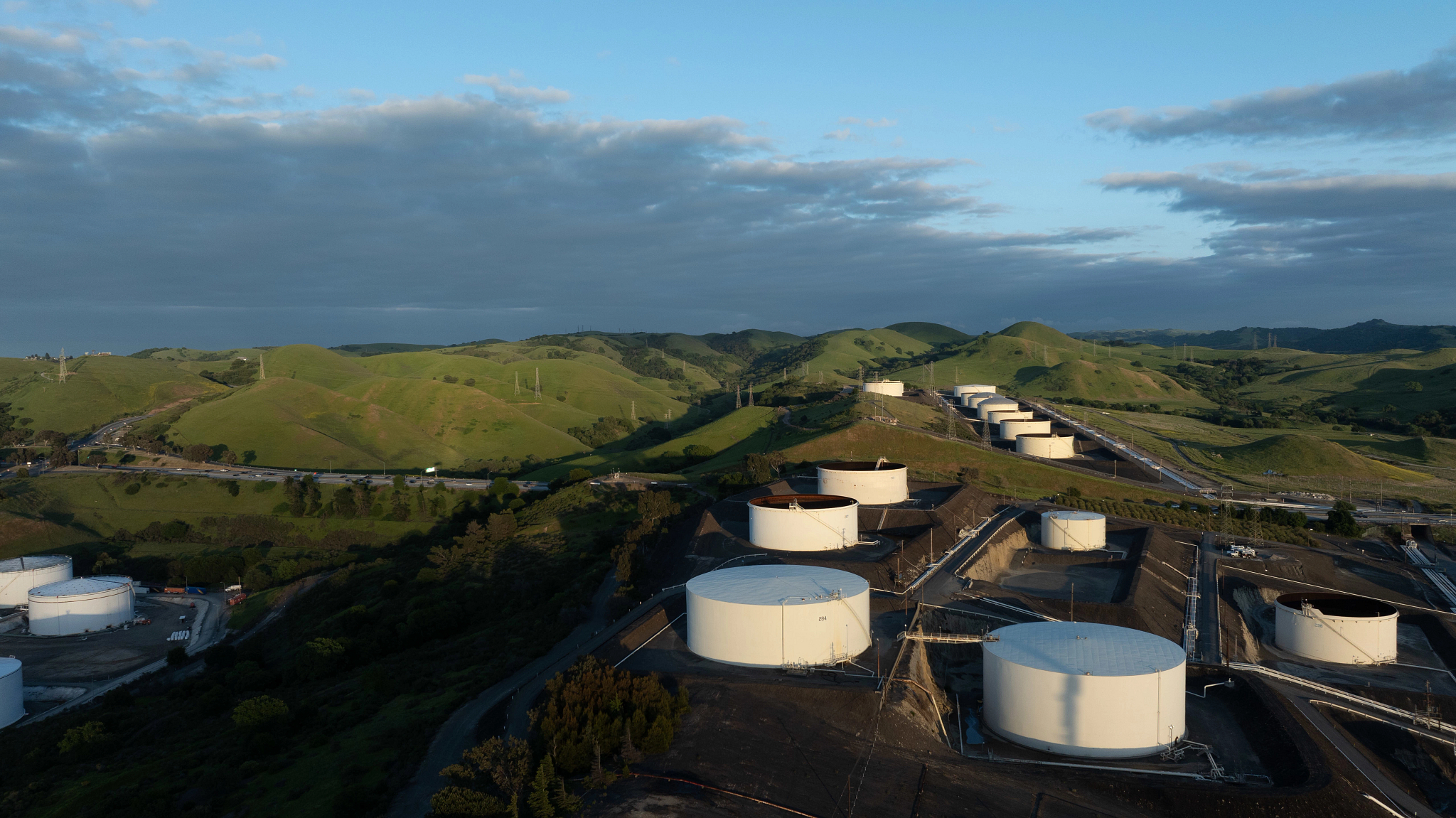 Aerial view of a refinery with large white storage tanks nestled among green rolling hills under a partly cloudy sky, illustrating the future of sustainable aviation fuel production.